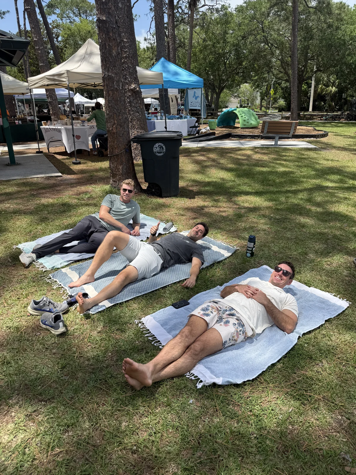 Three men relaxing on Salt Rags on the grass beside farmers-market tents.