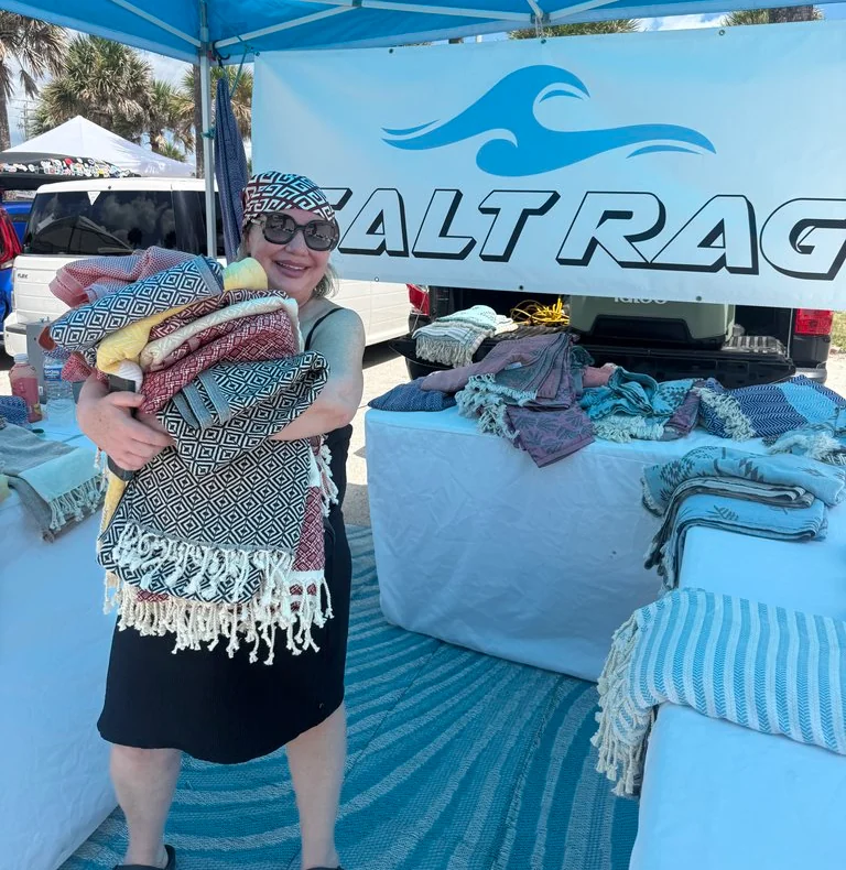 Smiling woman holding a large armful of patterned Salt Rags at the booth beneath the blue canopy.