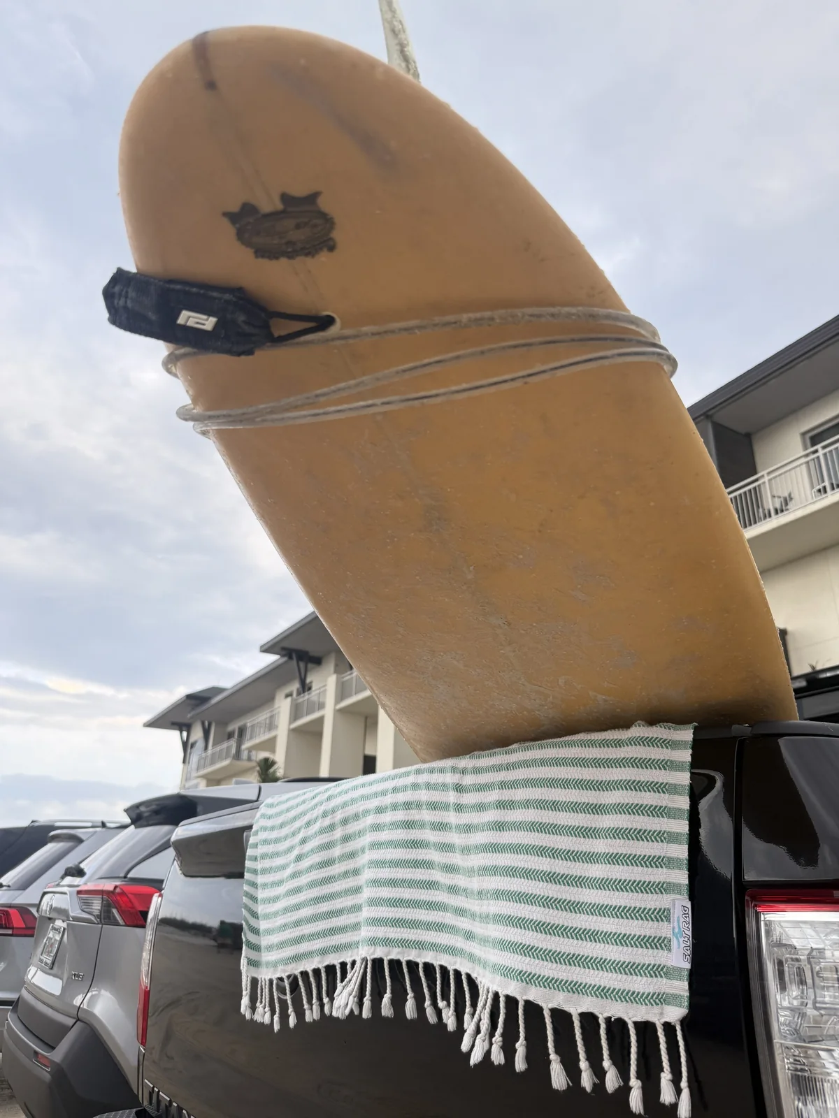 Green striped Salt Rag drying beneath a yellow surfboard on a parked vehicle.