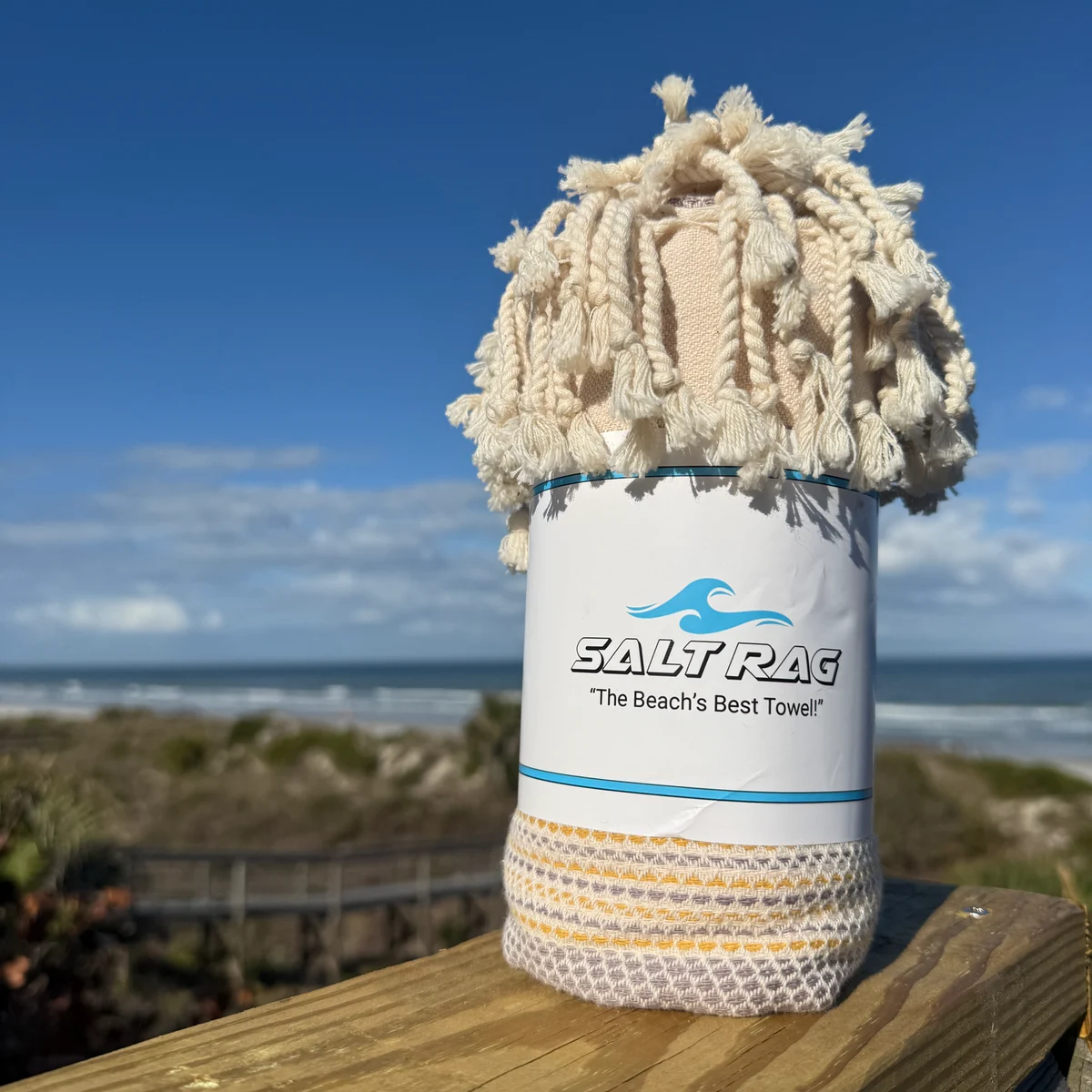 Pale yellow Salt Rag rolled on a wooden rail above the beach, with its fringe and band visible.