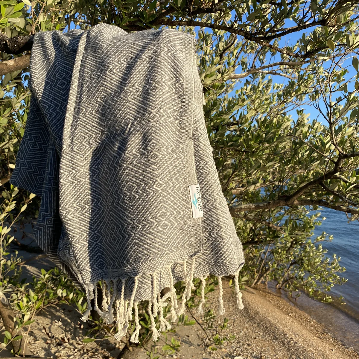 Gray geometric Salt Rag hanging from a coastal tree, showing the woven cotton, sewn cloth label and knotted fringe.
