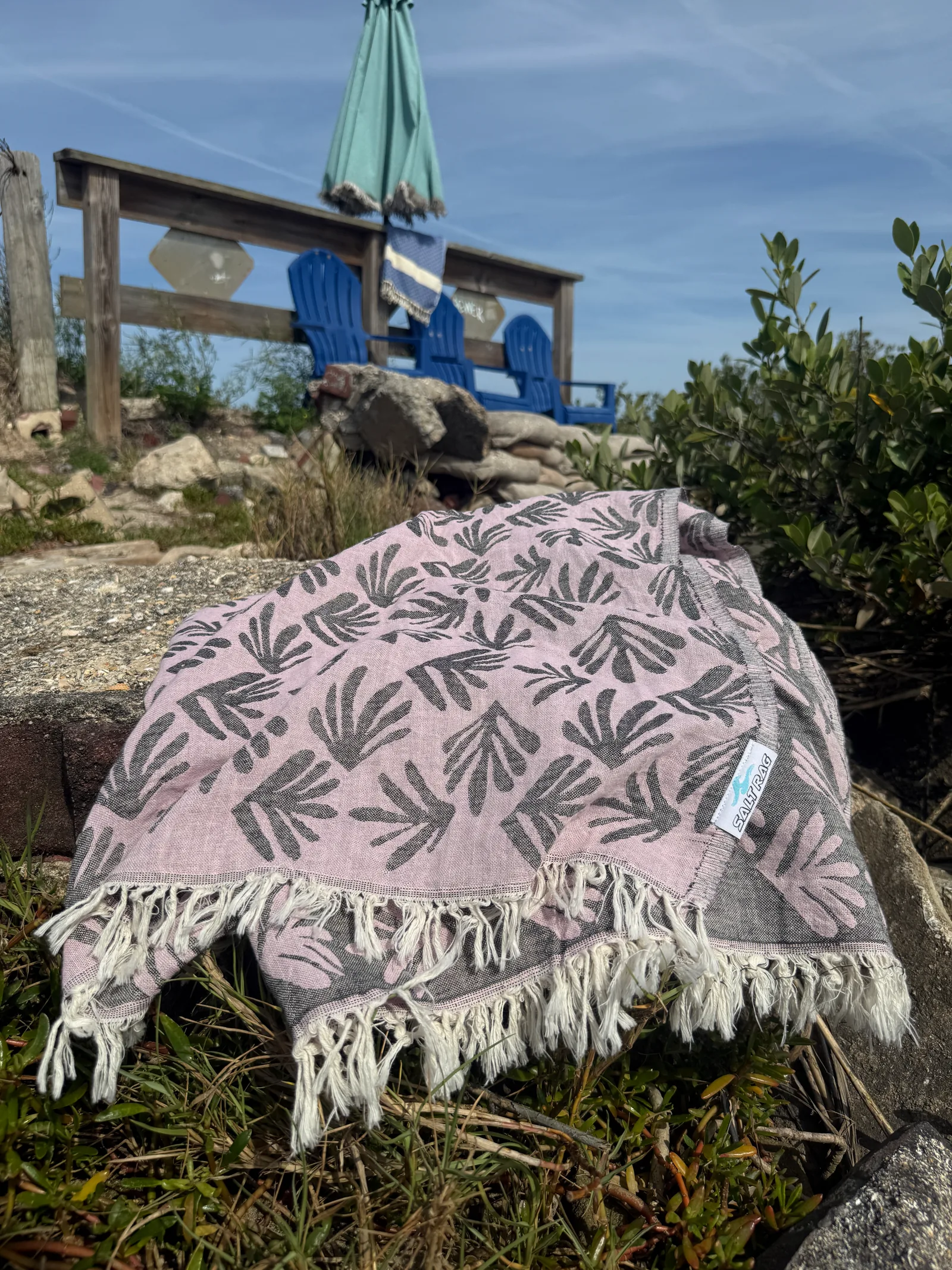 Pink leaf-patterned Salt Rag spread over coastal rocks near blue outdoor chairs.