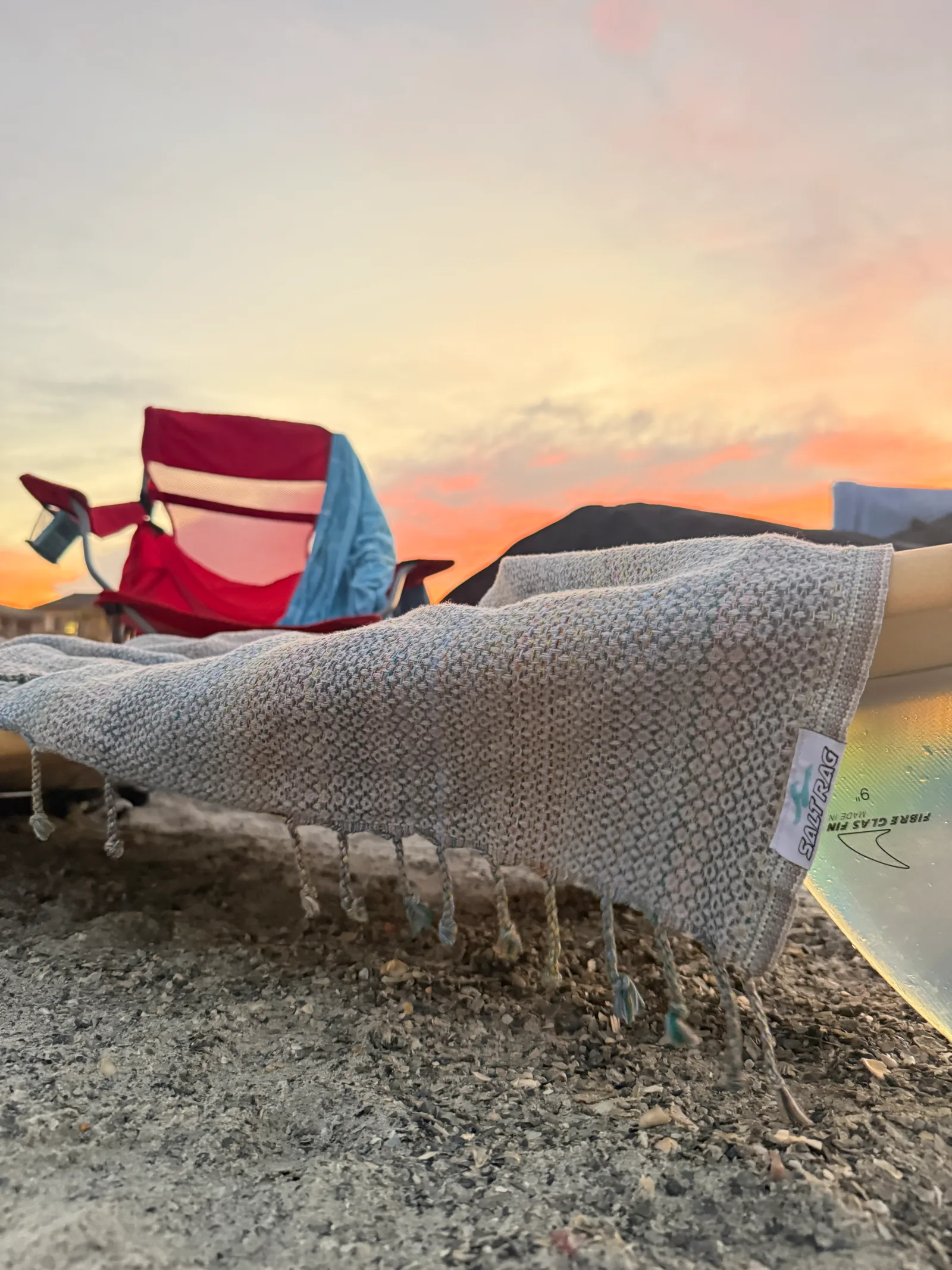 Gray woven Salt Rag draped over a surfboard on the beach, with a red chair and sunset behind.