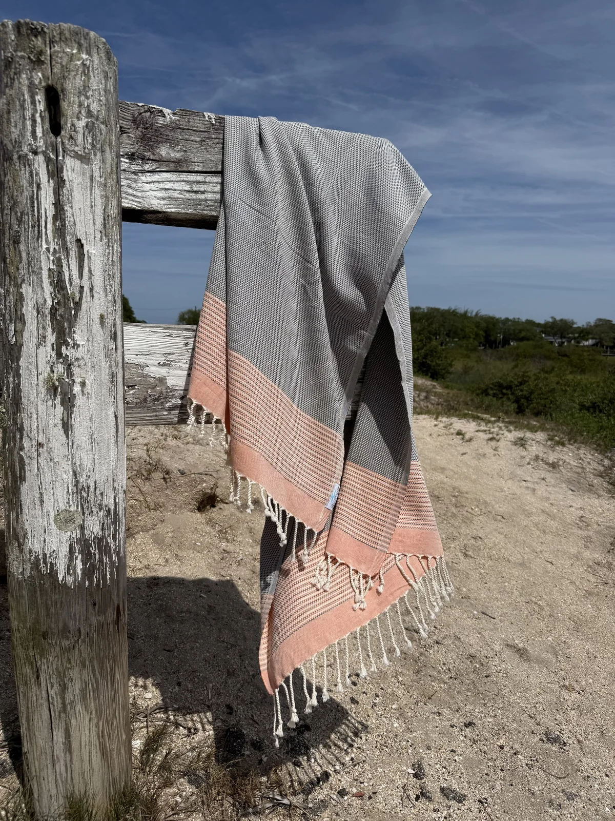 Gray and peach striped Salt Rag hanging from a weathered beach fence.