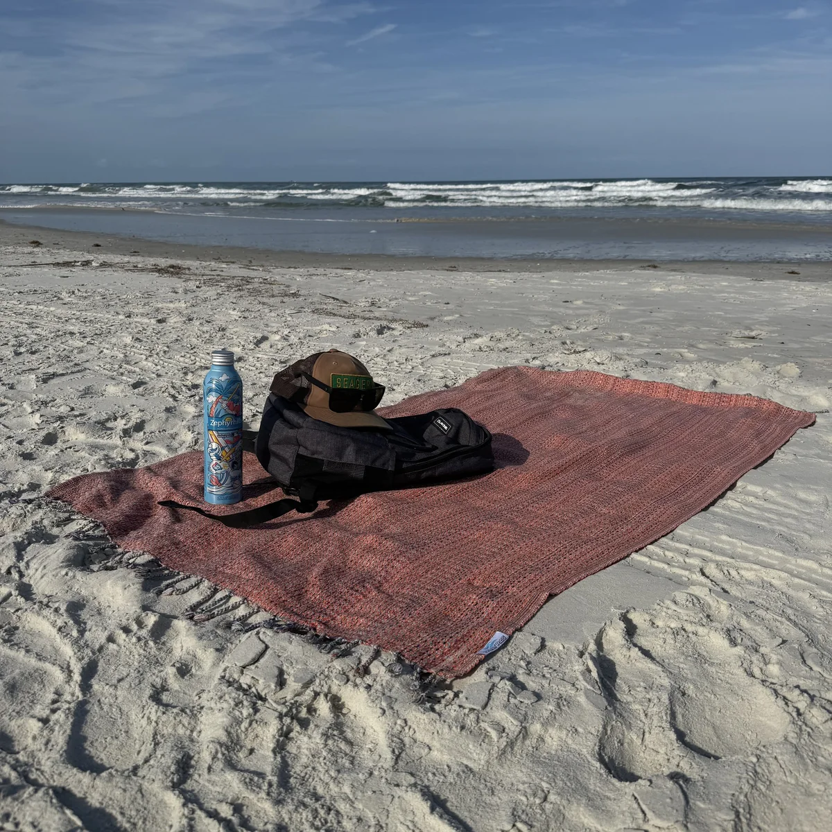 Beach flatlay with backpack, bottle and pink laid-out Rag.